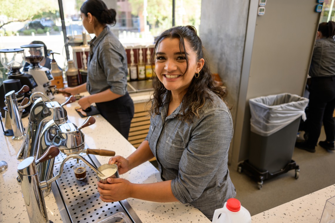 A young woman with darc curly hair froths milk at an espresso machine. 