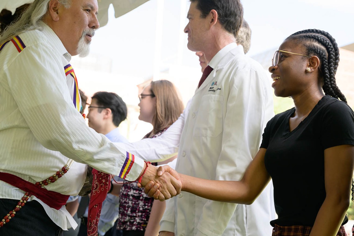 An older man in a Native American-style shirt shakes the hand of a young woman. 