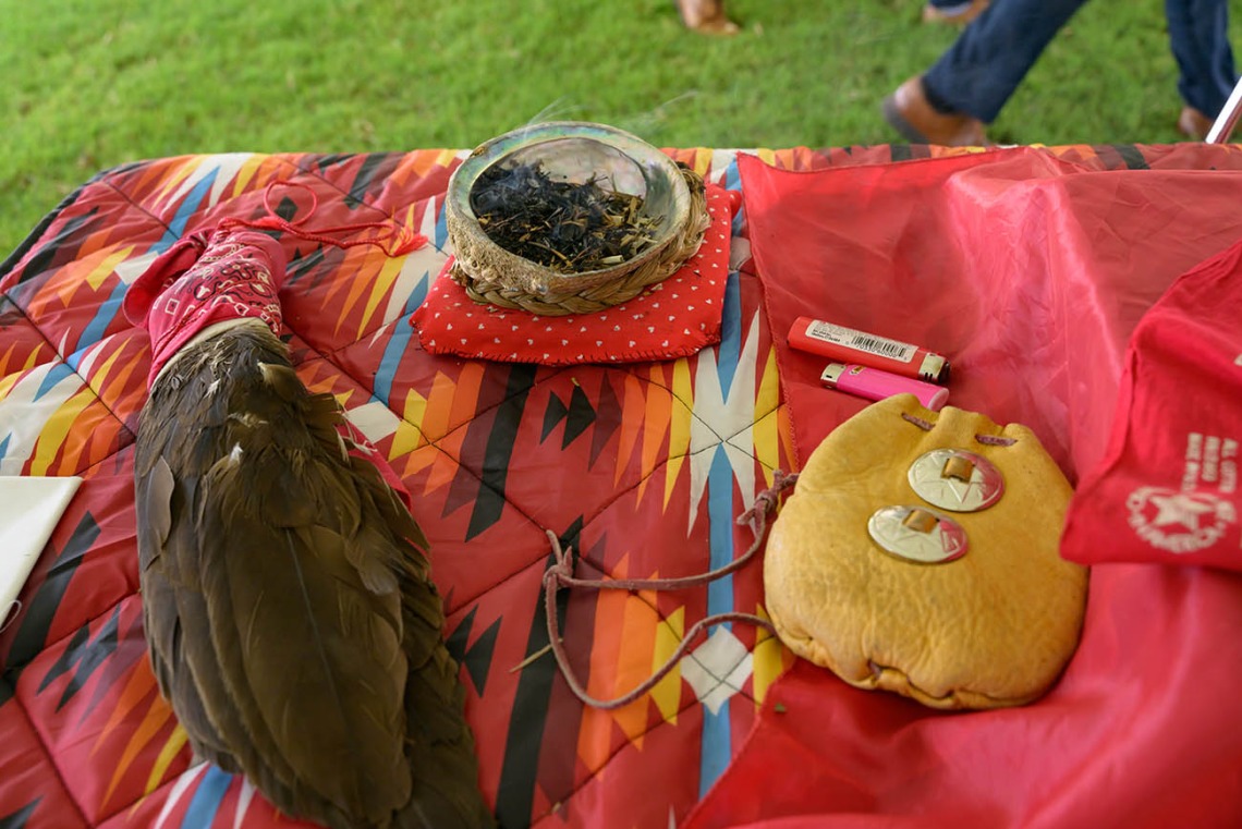 Table with red tablecloth and an eagle feather fan, an abalone shell a leather pouch sitting on it. 