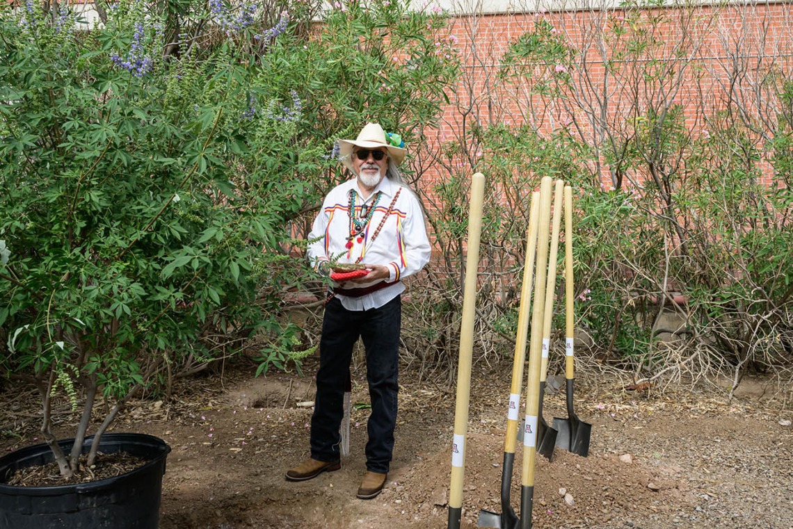 A man with a straw cowboy hat stands in a garden setting with several shovels stuck in the ground next to him and a tree in a big pot. 