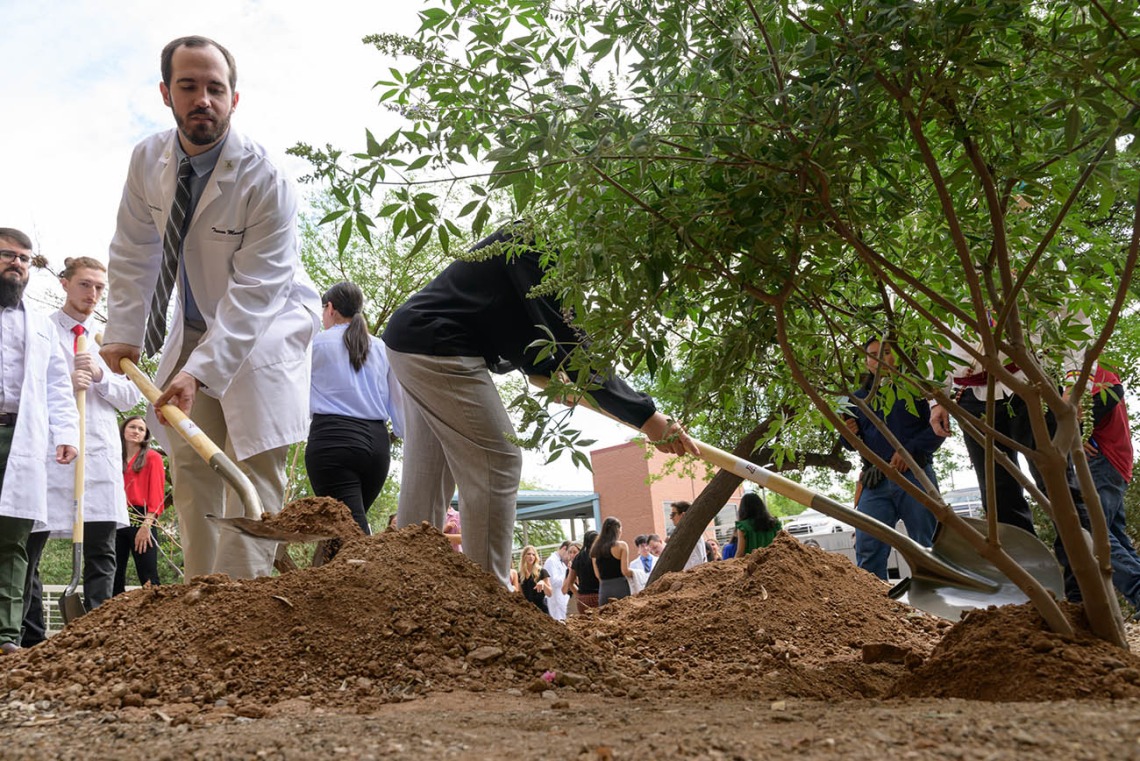 A man in a white medical coat shovels dirt into a hole with a tree in it. Several more medical students stand behind waiting for their turn.