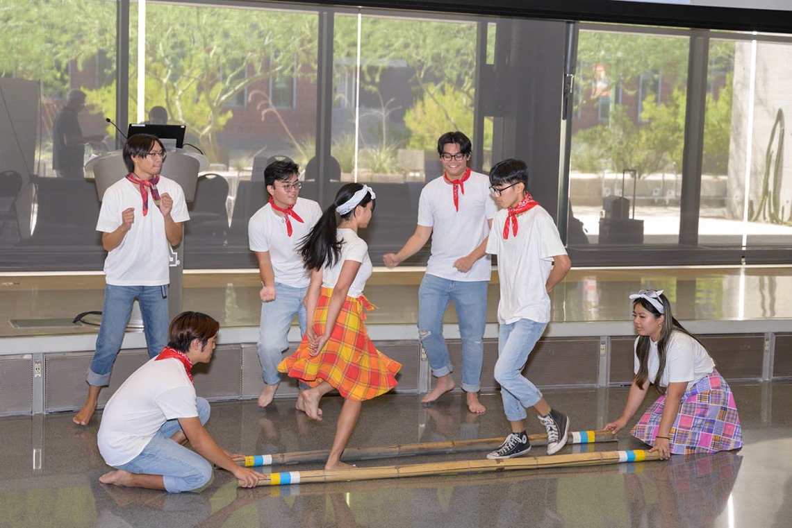 Seven students from the UArizona Filipino American Student Association perform a dance with two long wooden poles on the ground that they hop over.  