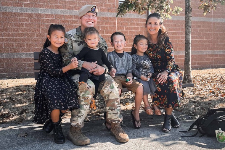 two parents and four children sitting on a bench. The father is wearing military camoflauge fatigues and a beret 