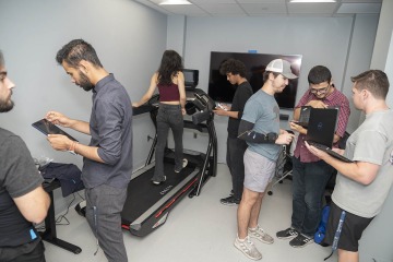 A group of students standing in a lab looking at computer screens, walking on a tread mill and looking at a tablet.