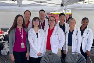 A diverse group of people (one female teacher and seven students) standing together and smiling under a tent. All but one person is wearing a white coat.