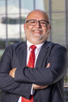 South Asian man with grey beard stands wearing a black suit, white shirt and red tie. His arms are folded and he is smiling.